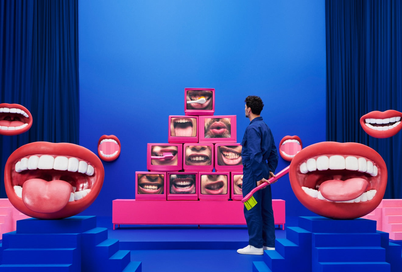 A person holding a giant toothbrush in front of a pyramid of screens showing mouths
